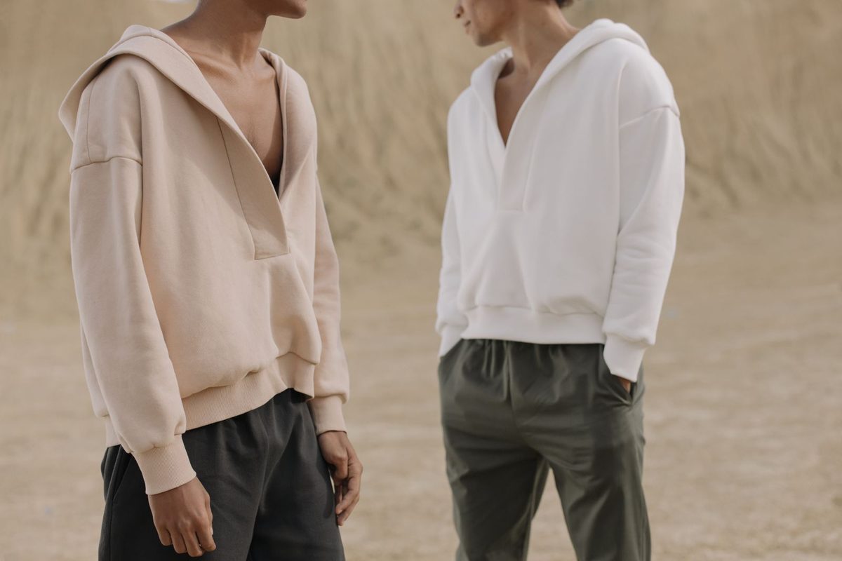 Two people in plain cream hooded sweatshirts and dark trousers standing on sand